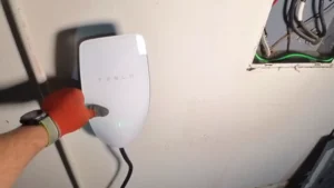 James installing a Tesla Gen 3 Wall Connector EV charger mounted on a wall, with an open electrical panel beside it showing circuit breakers and wiring setup