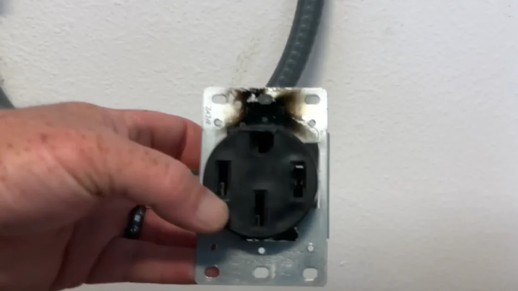 A close-up image showing James Ndungu holding a NEMA 14-50 electrical outlet that has been severely damaged by overheating. The plastic housing around the top terminals is melted and blackened with soot, clearly demonstrating the fire risk associated with using an improper outlet for the high, sustained power draw of electric vehicle charging.