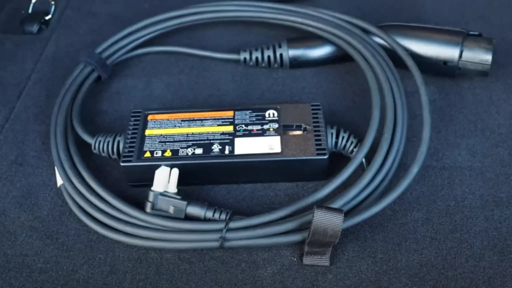 A close-up image highlights the Mopar Level 1 EV charger that comes standard with the 2025 Jeep Wrangler 4xe, neatly coiled and stored in the rear cargo area. The photo clearly displays the 120V power plug, power cable, compact charging unit, and J1772 connector. This charger plugs into any standard household outlet and delivers a full charge in approximately 12 hours. Its slim, portable design fits easily in the 4xe’s storage compartments, making it convenient for overnight charging at home or while traveling.