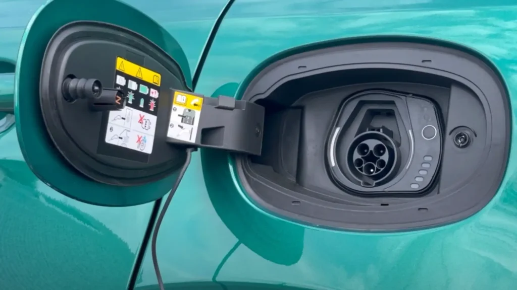 Close-up of Alfa Romeo Tonale’s SAE J1772 charging port with open flap, showing five-pin connector and black charging cable on a teal vehicle.