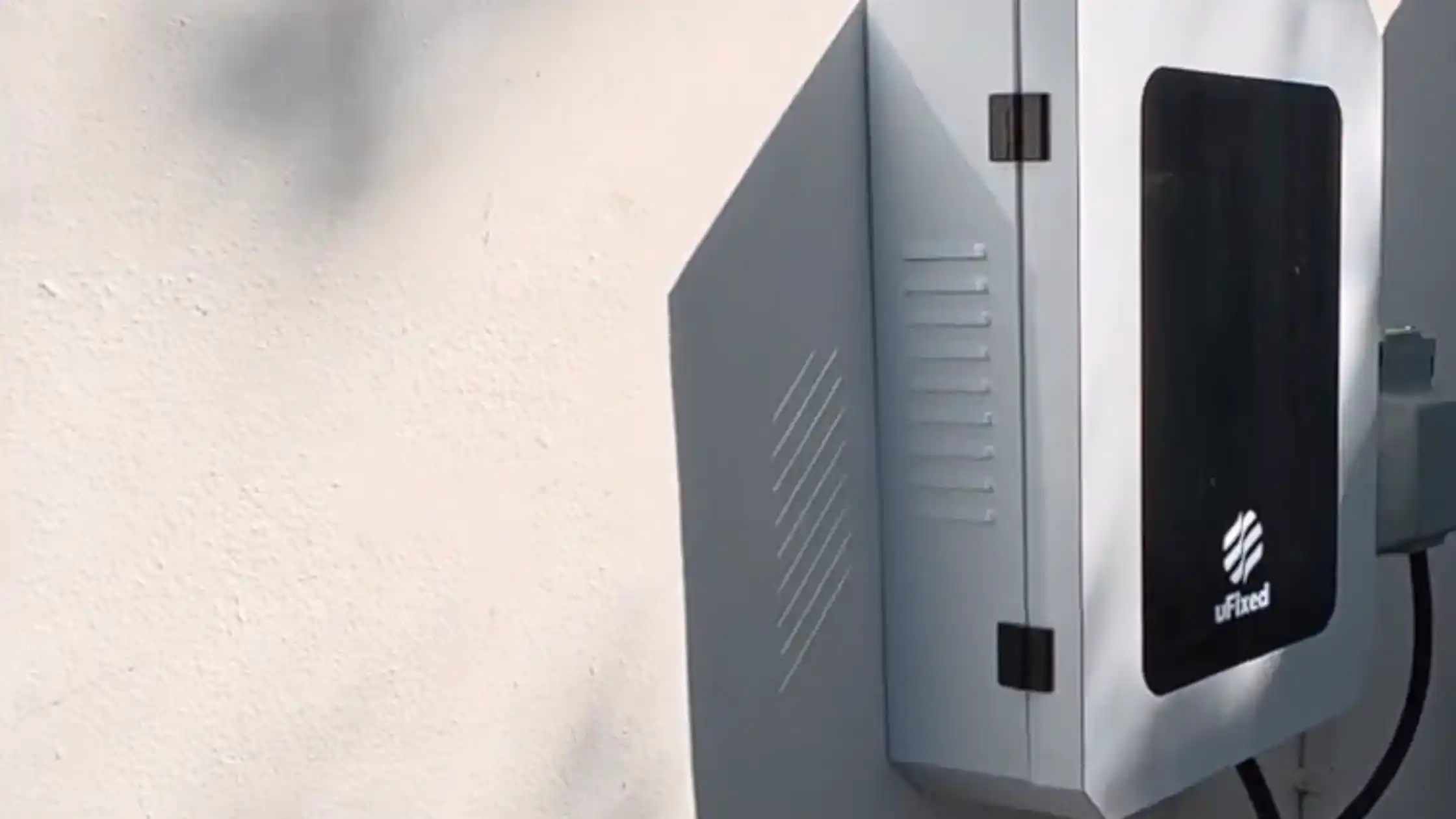 Light gray UFixed Outdoor EV Charger Enclosure mounted on textured exterior wall protecting a Tesla Wall Connector, with tinted dark front panel and ventilation vents.