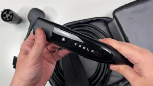 James shows a close-up of the Tesla Mobile Connector NACS handle with release button, with the coiled charging cable, storage bag, and plug adapters visible in the background.