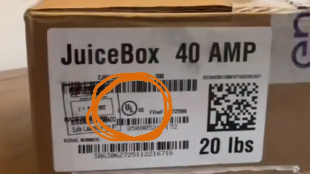 A close-up photograph of a white shipping or retail label affixed to a brown cardboard box for a JuiceBox 40 AMP Electric Vehicle (EV) charger. At the top of the label, the text "JuiceBox 40 AMP" is printed in large, bold black letters. In the center of the label, an orange hand-drawn circle highlights the Underwriters Laboratories (UL) certification mark, which consists of the letters "UL" inside a circle with a small "US" printed to its right, indicating the product is certified for use in the United States. To the right of the UL mark is the text "File# E472596" (a unique identifier for the manufacturer's certification record). The label also features a large QR code on the right side, a "20 lbs" weight specification at the bottom right, and various barcodes, serial numbers, and smaller manufacturing details across the surface.