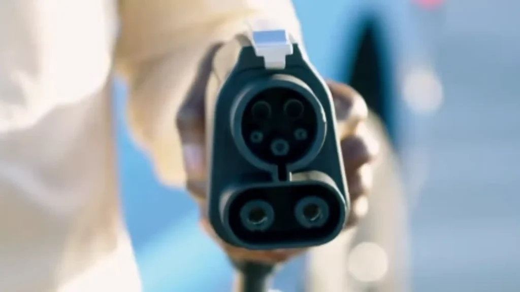 A close-up of a person holding a black CCS Combo 1 electric vehicle charging plug, showing the top five-pin circular interface and the two large DC pins at the bottom.