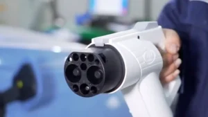 James is holding a white and black GB/T DC fast-charging connector, showing the distinctive nine-pin layout used by electric vehicles in China.