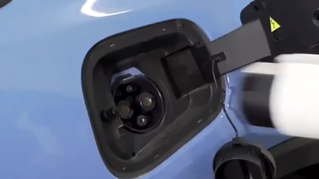 A close-up of a blue electric vehicle with its charging door open, revealing a black GB/T DC charging inlet as a white charger is brought toward it.