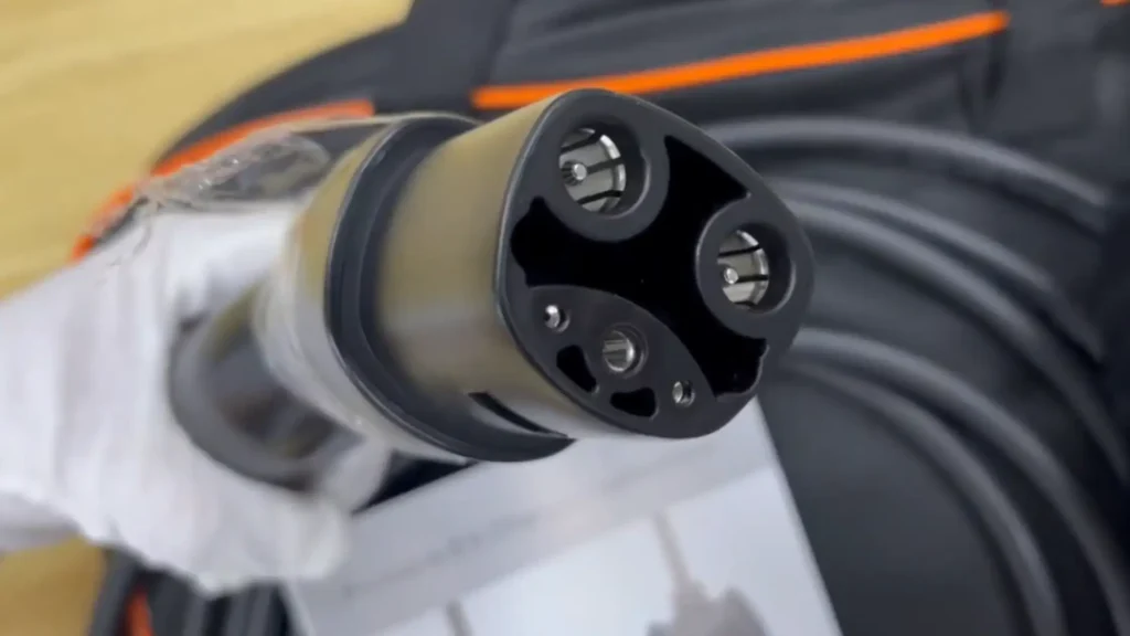 James is wearing a white glove while holding a Tesla NACS electric vehicle charging connector with visible charging pins