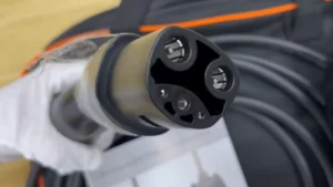 James is wearing a white glove while holding a Tesla NACS electric vehicle charging connector with visible charging pins