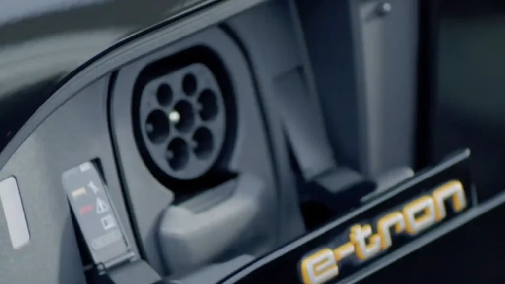A close-up view of the open charging port on an Audi e-tron, showing the seven-pin Type 2 Mennekes inlet and the "e-tron" branding on the motorized door.