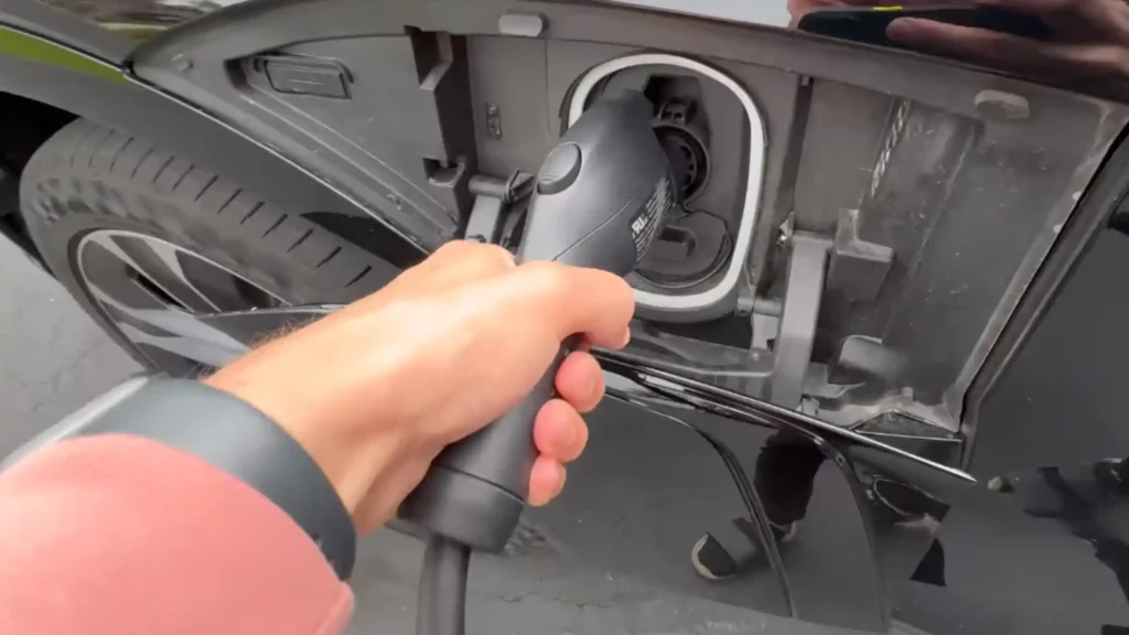A first-person perspective shot of James holding a black EV charging handle, preparing to plug it into the side-mounted charging port of a 2025 Chevrolet Blazer EV RS in high-gloss black. The vehicle's aerodynamic RS-trim wheel and tire are partially visible in the background.